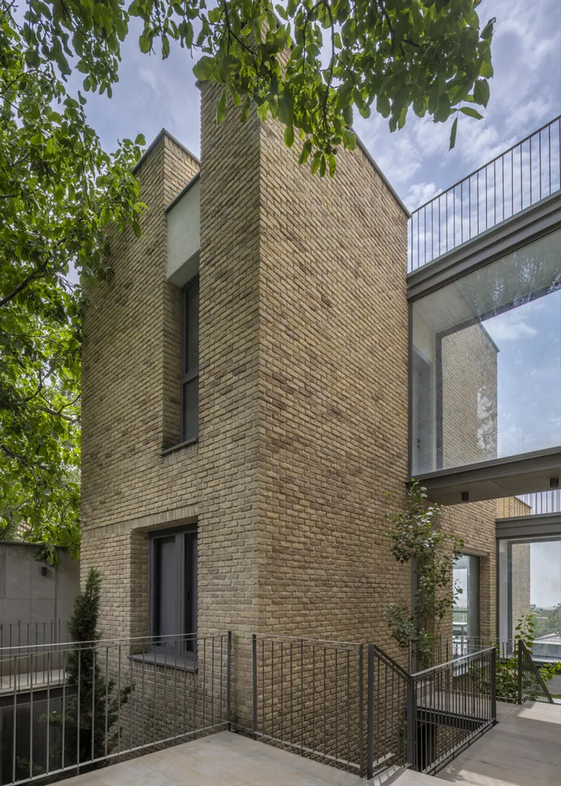 Buff brick tower volume with narrow vertical window slot framed by overhanging tree branches