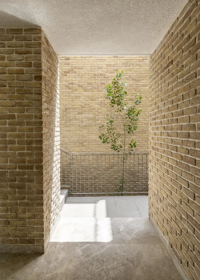 Narrow brick-walled courtyard passage with young tree behind metal railing in dappled sunlight