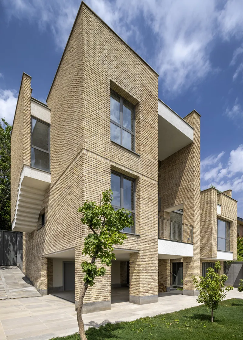 Cream brick facade with stacked volumes and white balconies framed by young trees in a sunny courtyard