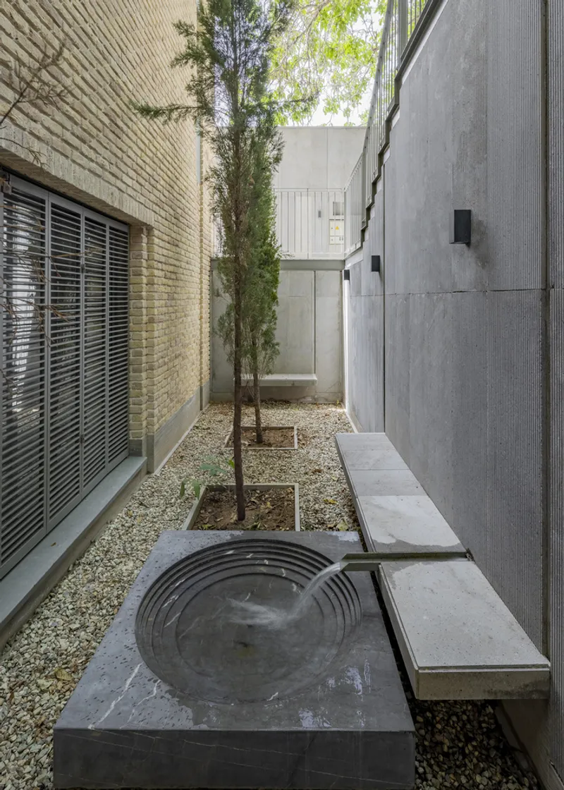 Narrow side courtyard with circular stone water feature between textured concrete wall and brick facade