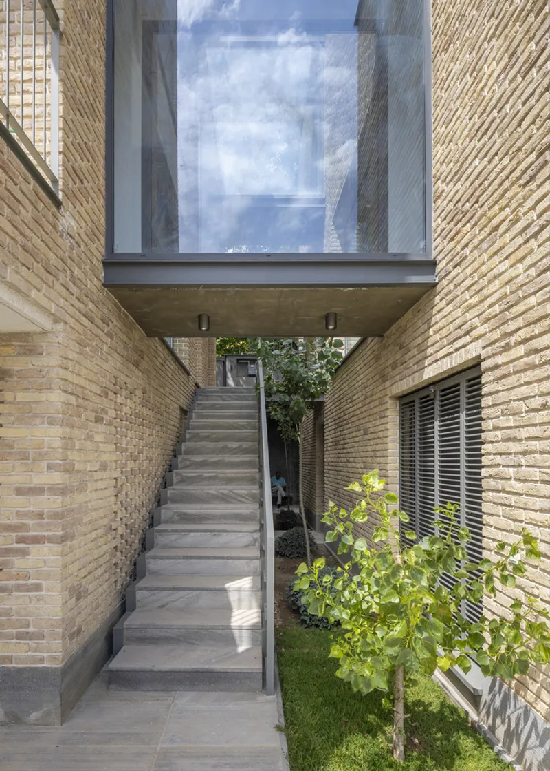 Exterior concrete staircase beneath glazed bridge connecting buff brick volumes with planted bed alongside