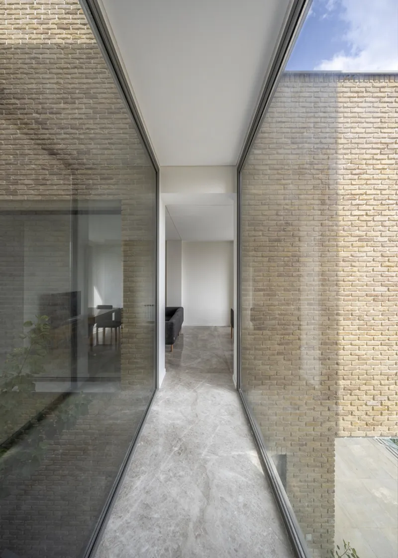 Glass-walled passageway between brick walls reflecting interior furnishings under an open sky
