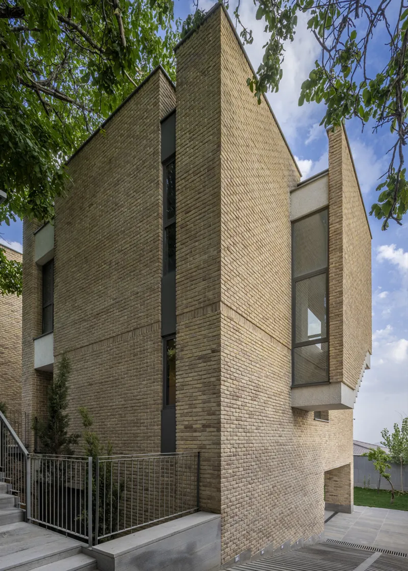 Angled brick facade with narrow vertical windows and steel railing beneath leafy tree canopy