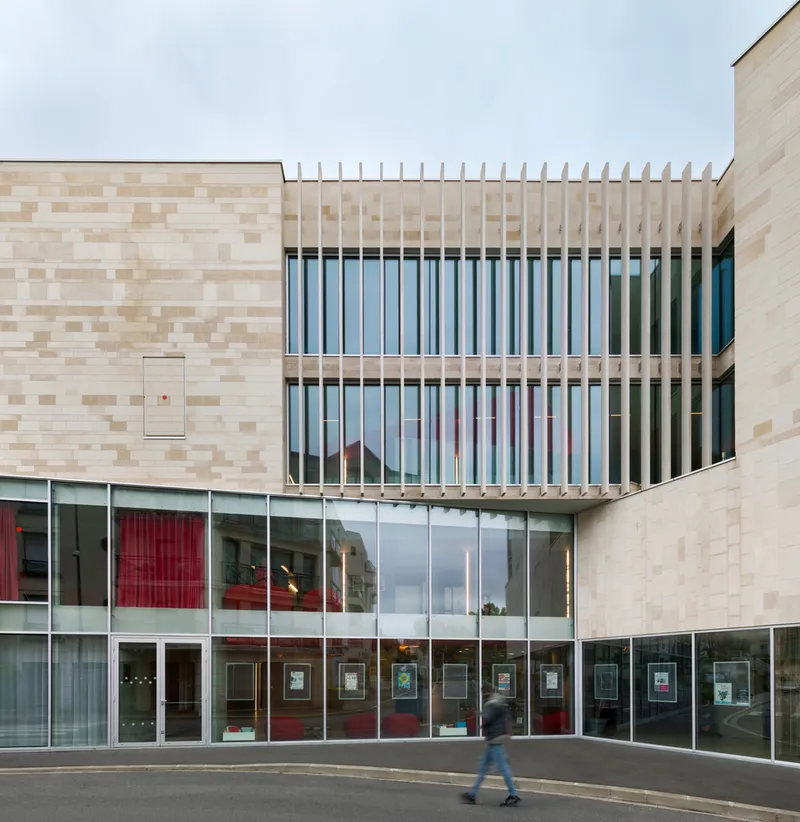 Street facade with variegated brick walls, glazed ground floor and vertical timber brise-soleil as a pedestrian passes
