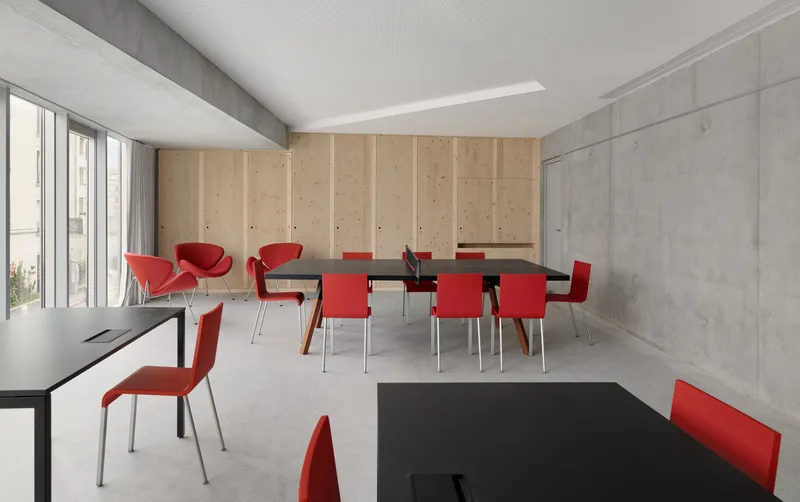 Meeting room with exposed concrete walls, plywood cabinetry, black tables and red upholstered chairs