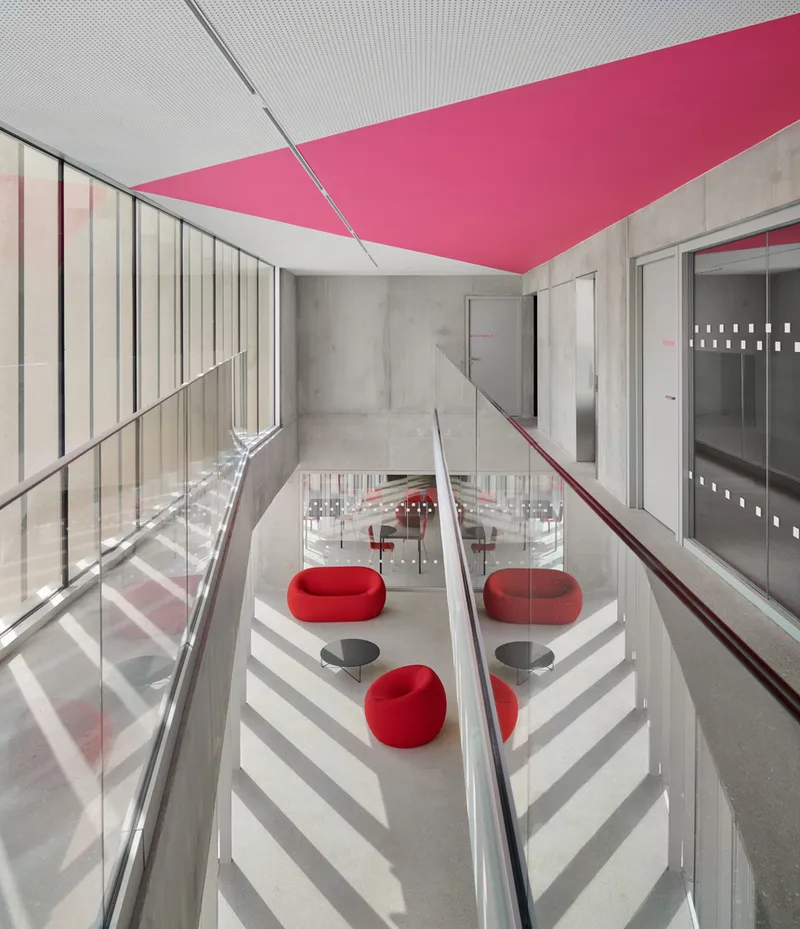 Upper level circulation space with pink diagonal ceiling element and glass balustrade overlooking seating below