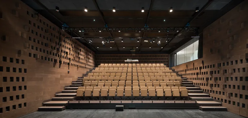 Auditorium with timber seating risers and perforated brick sidewalls beneath an exposed black ceiling grid