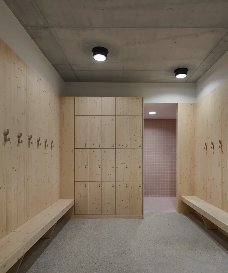 Changing room with light wood lockers, benches and concrete ceiling with glimpse of pink tiled space beyond