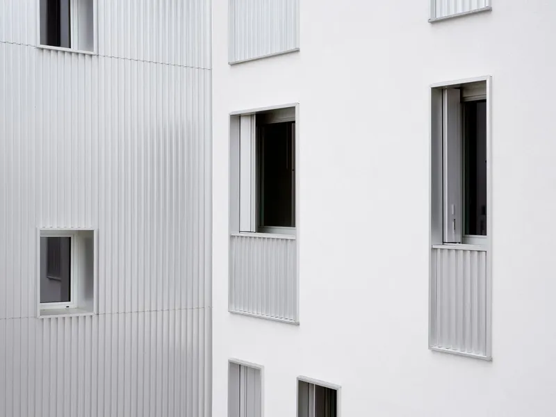 Close-up of windows and corrugated panels between white rendered facades under overcast sky
