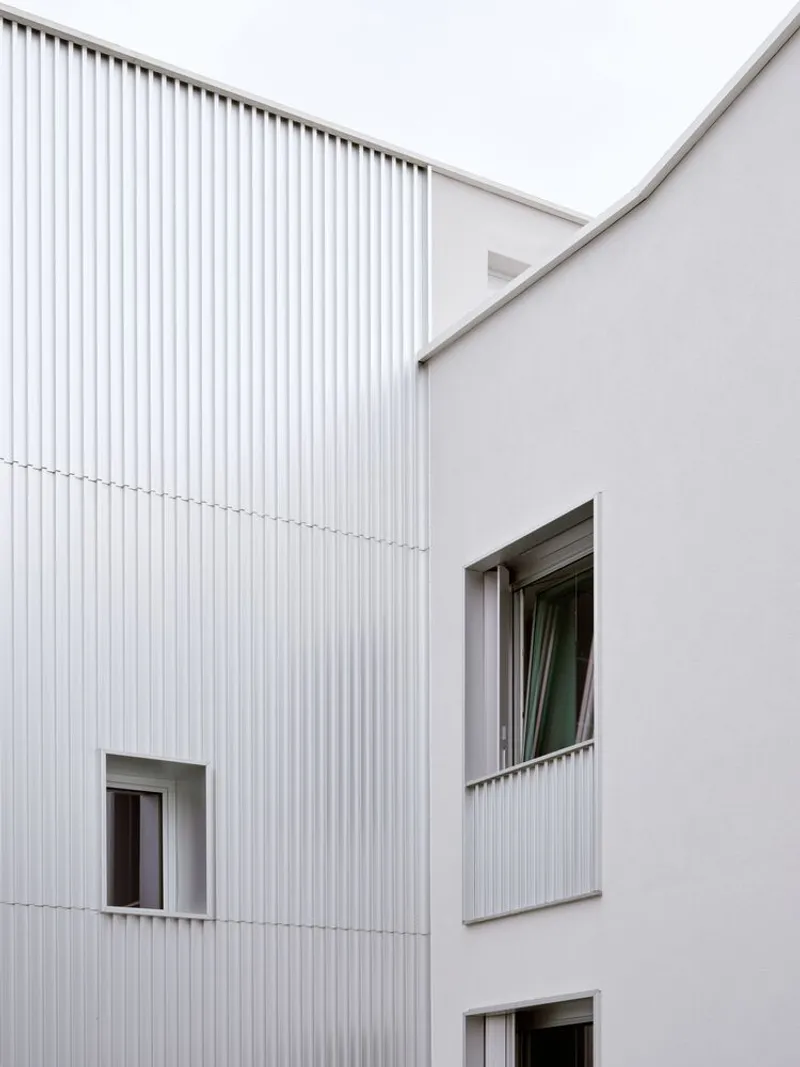 White corrugated metal and smooth plaster facade with scattered window openings under overcast sky