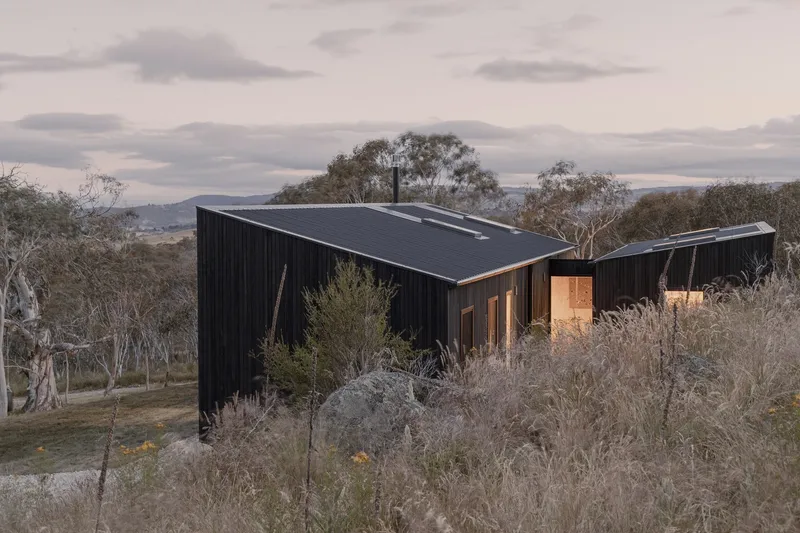 Black corrugated metal volumes nestled into a hillside with native grasses under an overcast dusk sky