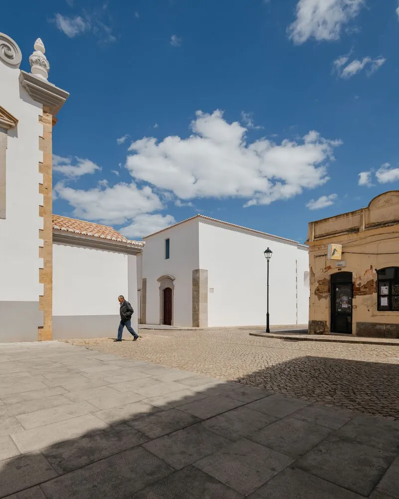 Cobblestone plaza with white plastered chapel and stone-trimmed corner building under a blue sky with clouds