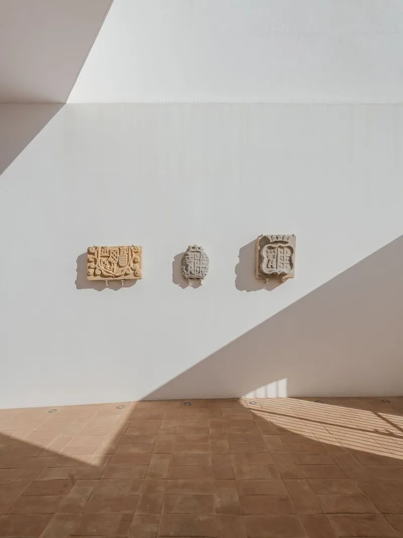Interior gallery wall with three carved stone crests mounted above terracotta tile flooring in angled sunlight