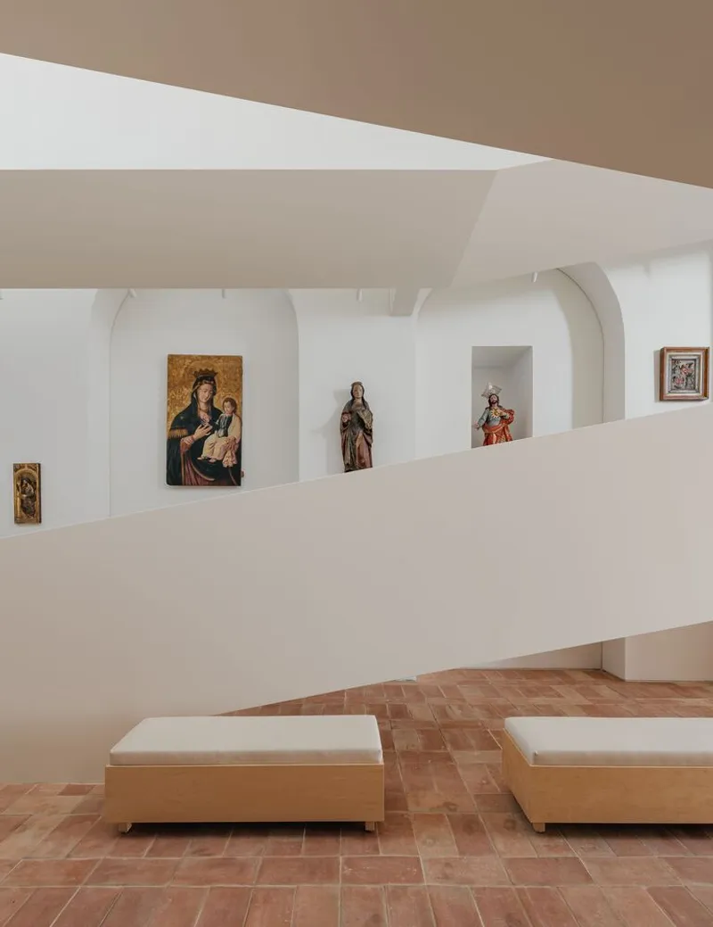 Gallery wall with arched niches displaying religious artworks above upholstered benches on terracotta tile flooring