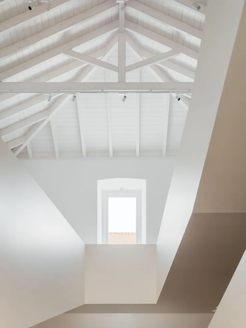 Interior gallery space with exposed white-painted timber roof trusses and clerestory opening framing tiled roofscape beyond