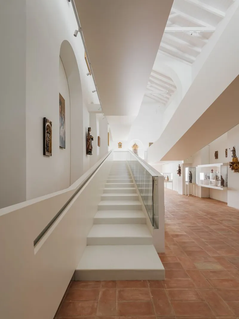 White staircase with glass balustrade ascending between gallery levels under exposed timber ceiling structure