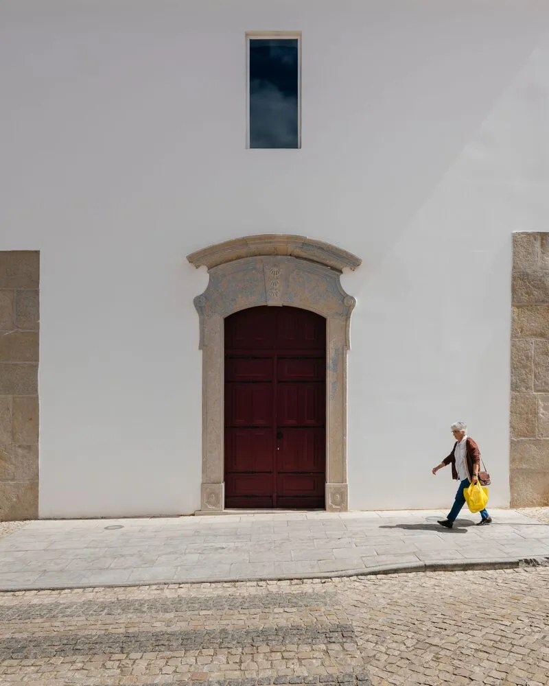 White stucco facade with stone-framed wooden door and narrow window as a pedestrian passes