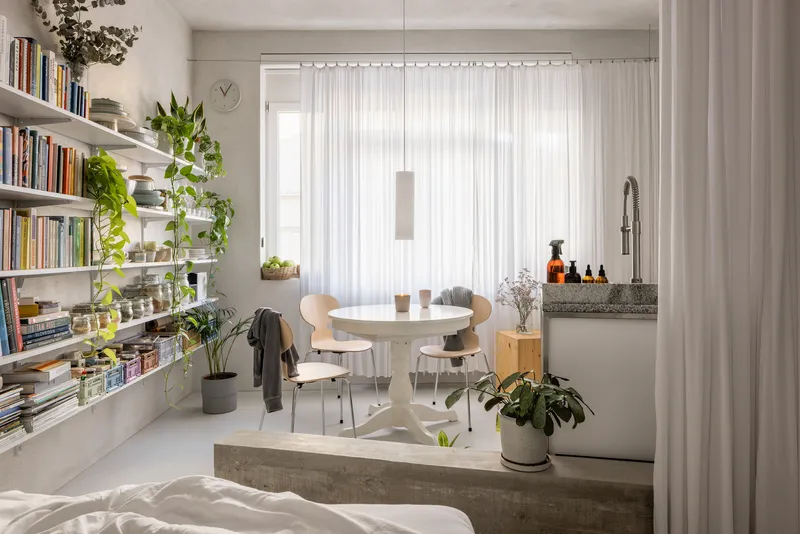 Open plan living space with white shelving, round dining table, and sheer curtains filtering daylight