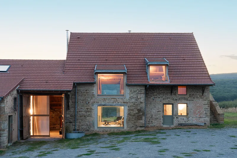 Stone barn with illuminated window openings and clay tile roof at dusk