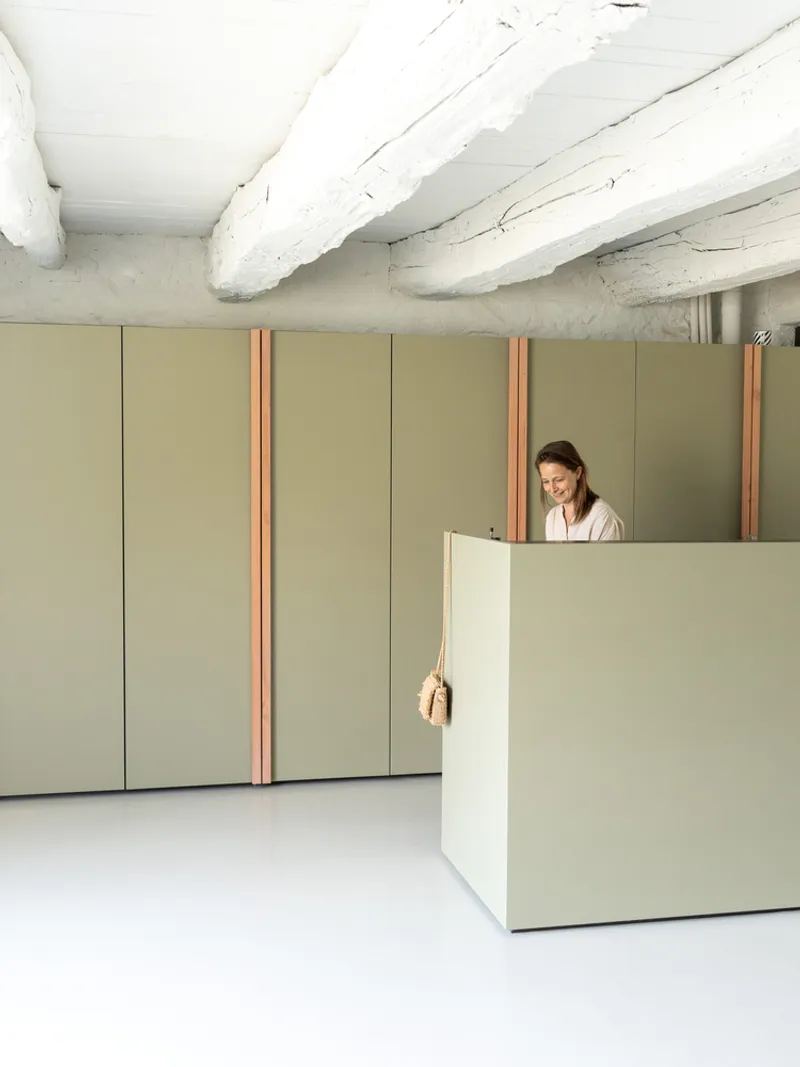 Pale green cabinetry with copper trim beneath whitewashed ceiling beams and a woman at the desk
