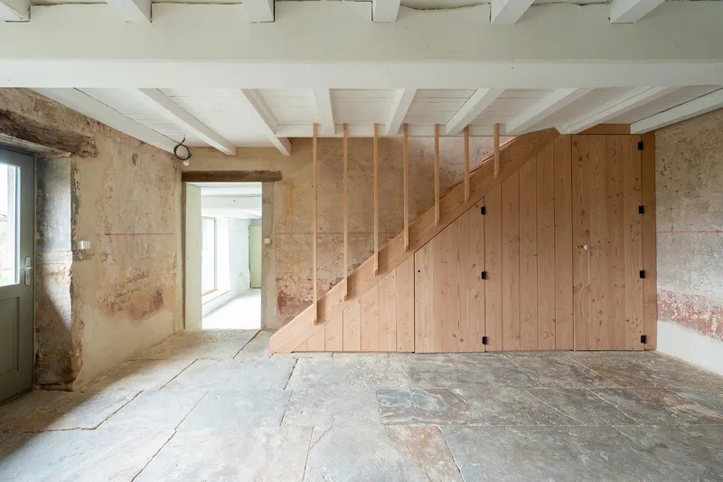 Timber staircase and storage volume against weathered stone walls with painted white ceiling joists