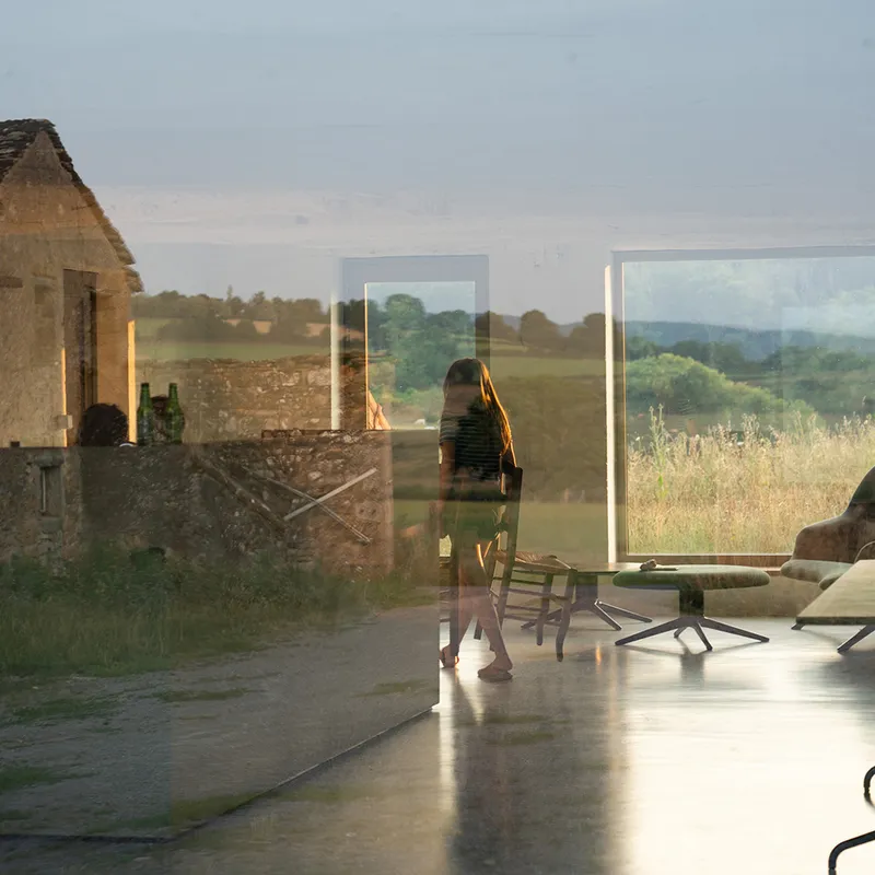 Interior view through floor-to-ceiling glass walls framing a figure and the rural landscape at golden hour