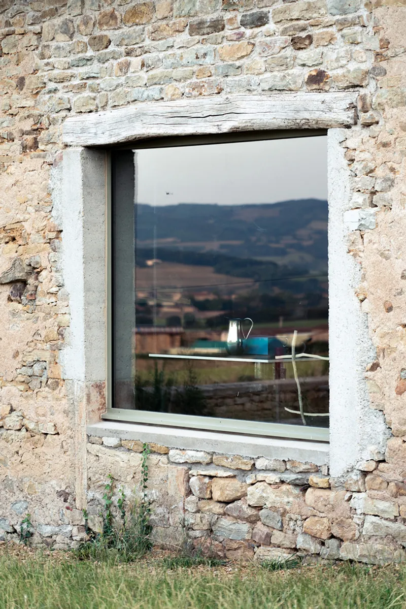 Window opening in weathered stone wall with salvaged timber lintel and white-framed glazing reflecting the hillside
