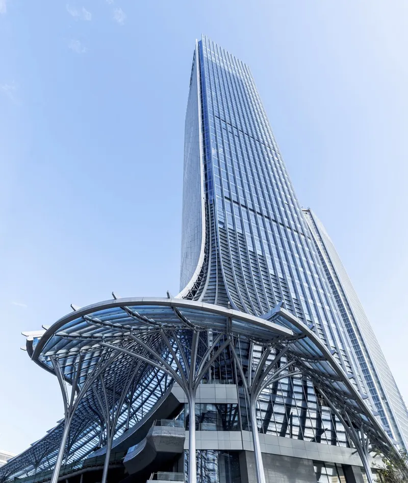 Street-level view of tapered glass tower rising from branching steel canopy base in daylight