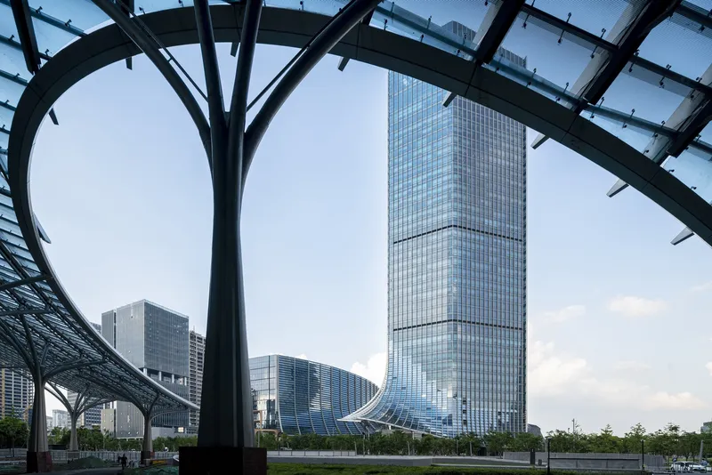 View through steel canopy structure toward glass tower and adjacent buildings under overcast sky