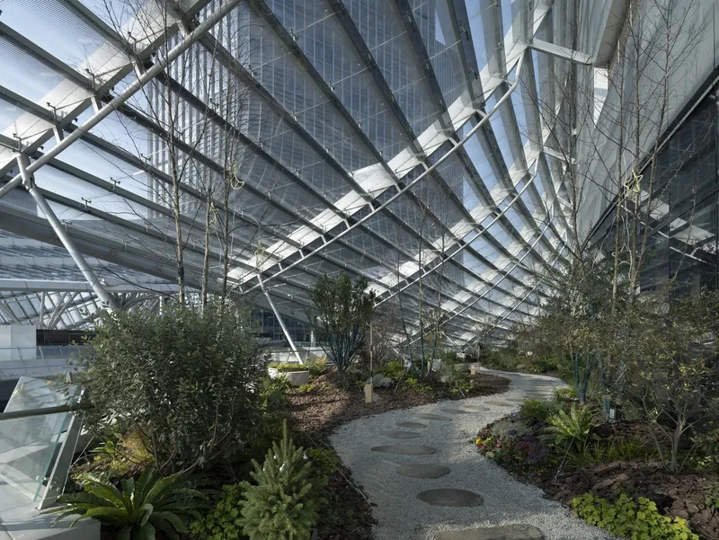 Enclosed garden path with planted beds and trees beneath diagonal steel and glass roof structure