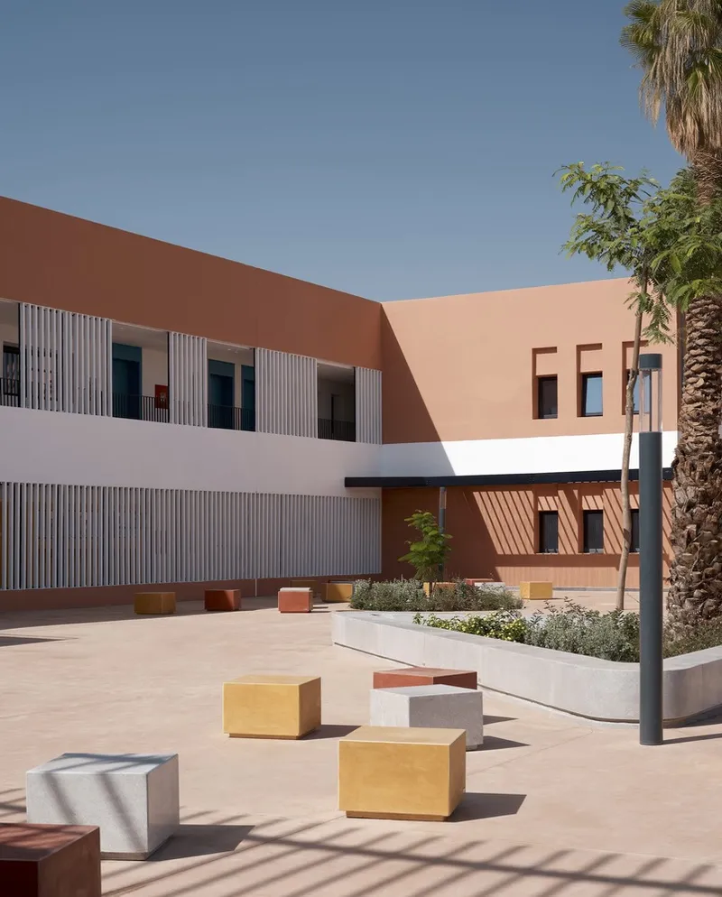 Paved courtyard with colored concrete seating cubes, raised planters, and vertical screens on the facade under clear blue sky