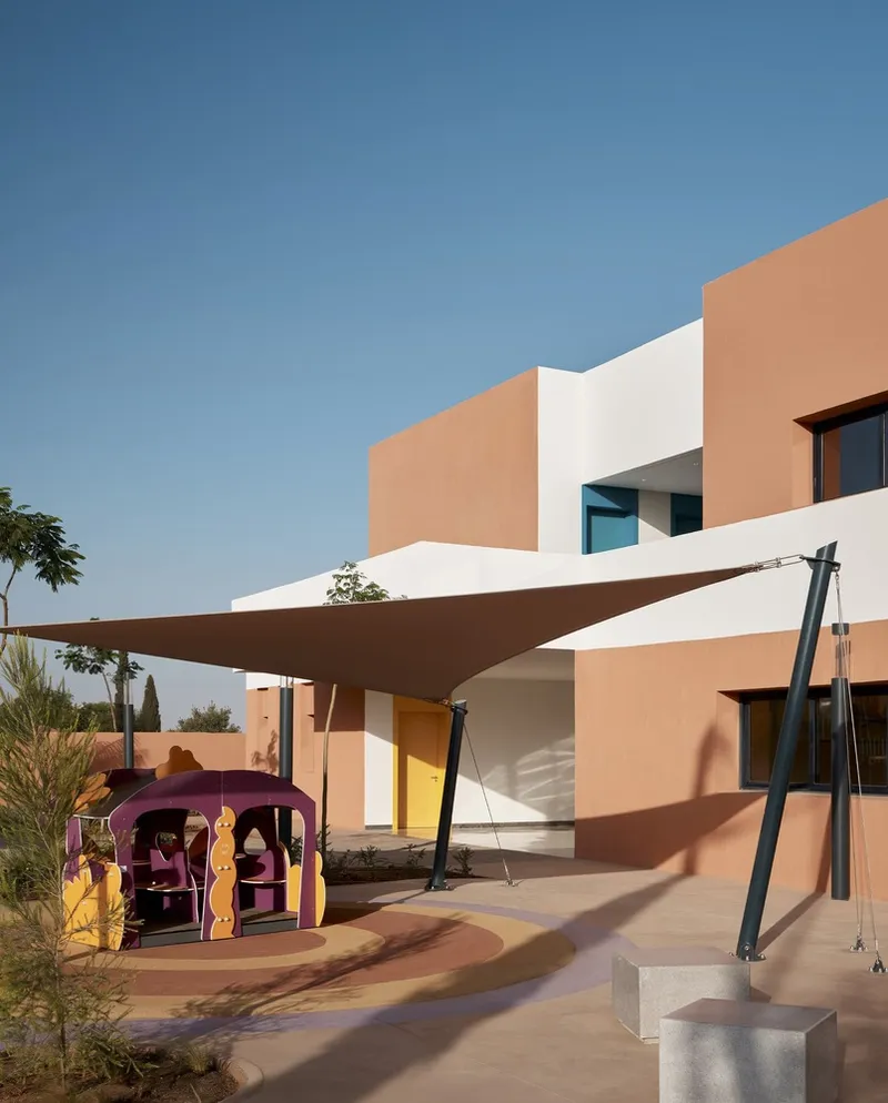 Outdoor play area with purple play structure, swing set, and tensile fabric shade canopy beneath terracotta facades