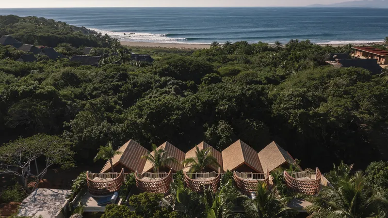Distant view of the clustered pavilions with pitched roofs and perforated facades facing the ocean through forest canopy