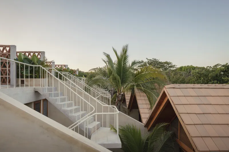 External staircase with white tubular railings ascending past pitched timber roofs and perforated brick screens at sunset