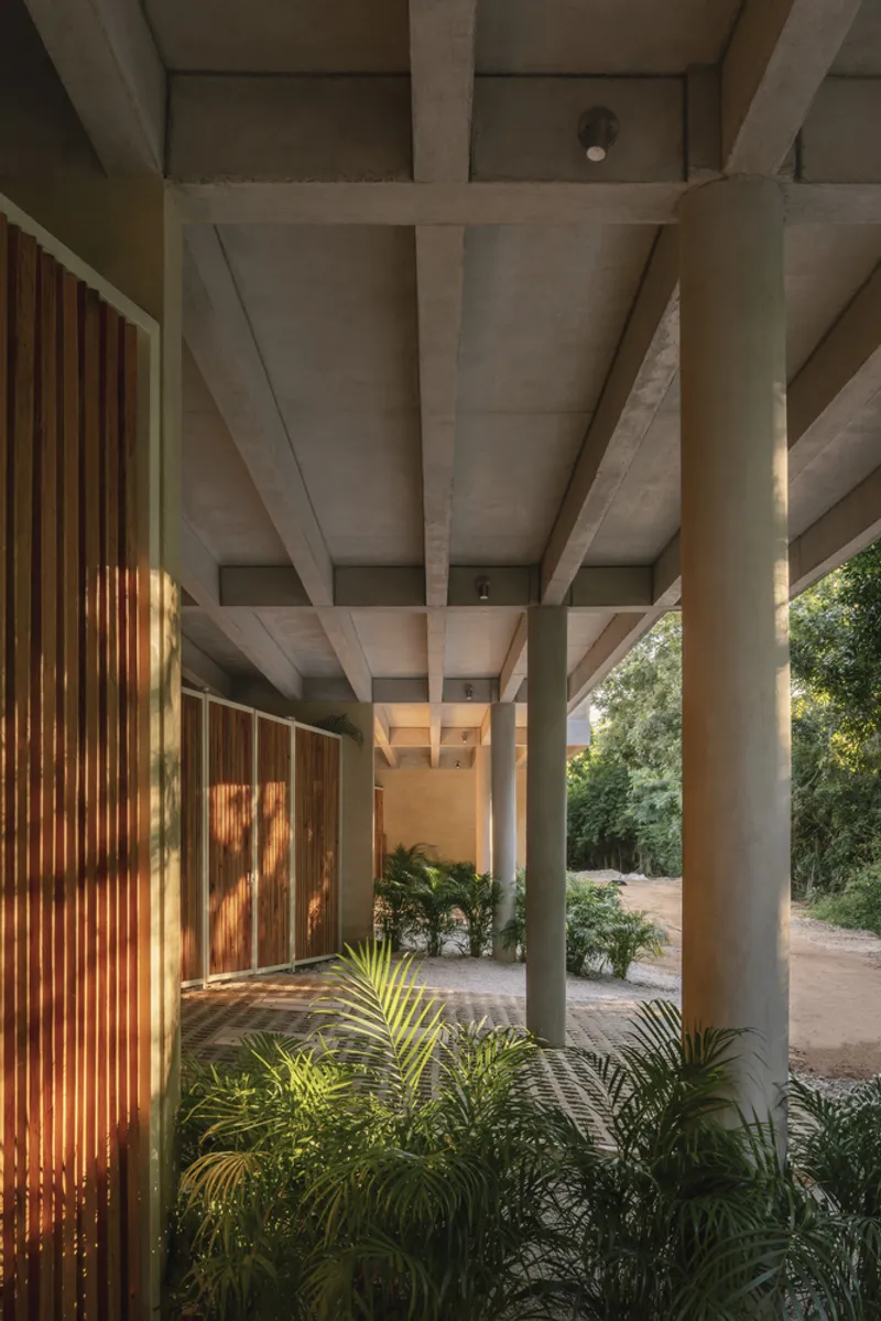 Covered walkway with exposed concrete beams and cylindrical columns framing vertical timber slat screens and potted palms