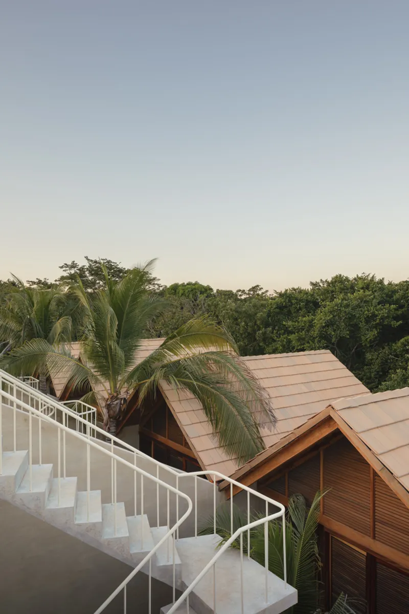 White curved staircase descending from a rooftop terrace past palm trees and pitched timber roofs at dusk