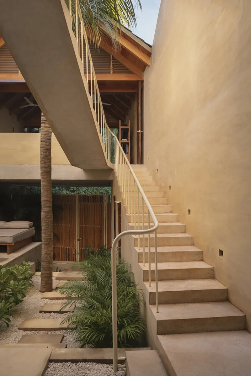 Exterior staircase with pale stone treads and curved metal railing rising beside planted courtyard with palm