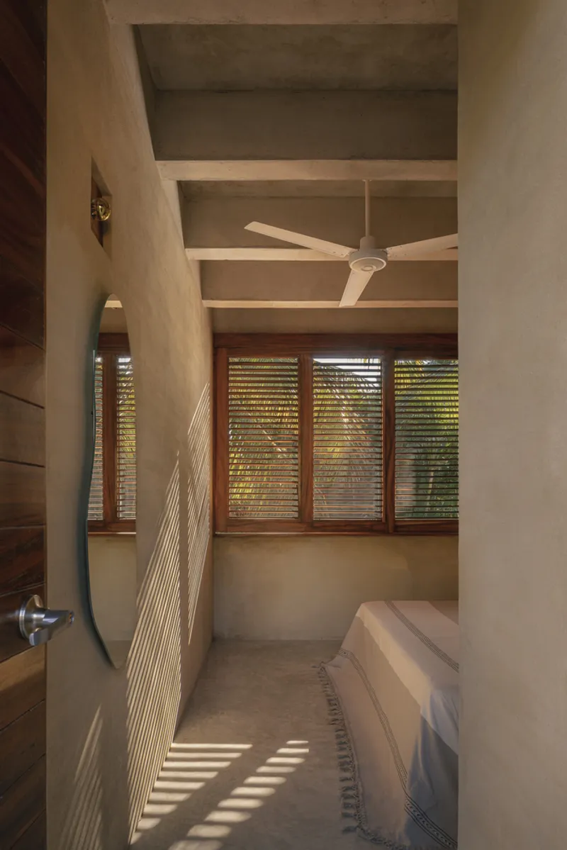 Bedroom with exposed concrete beam ceiling and timber louvered windows filtering afternoon sunlight across floor