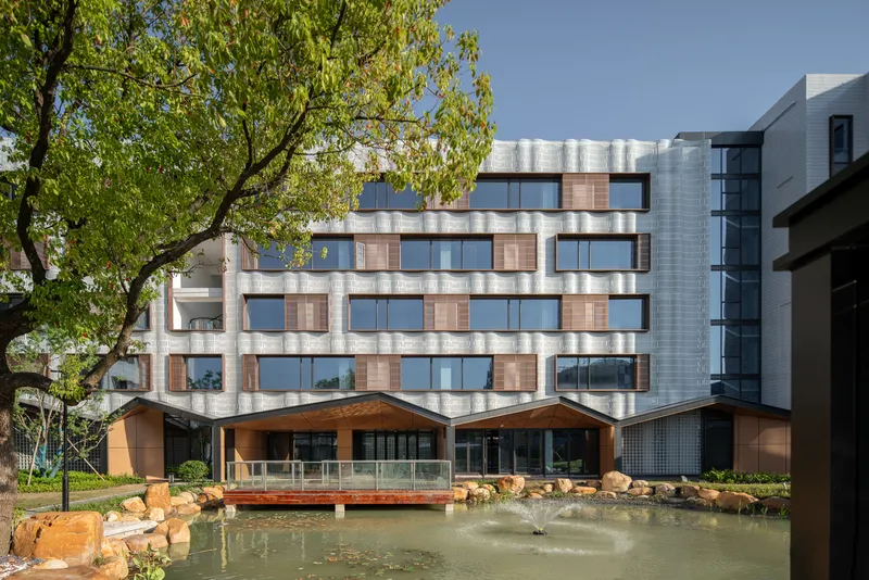 Facade with alternating glass windows and timber shutters overlooks a pond edged with rock boulders