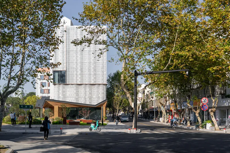 Stacked volume with ribbed metal facade rising above a timber-framed base and tree-lined street
