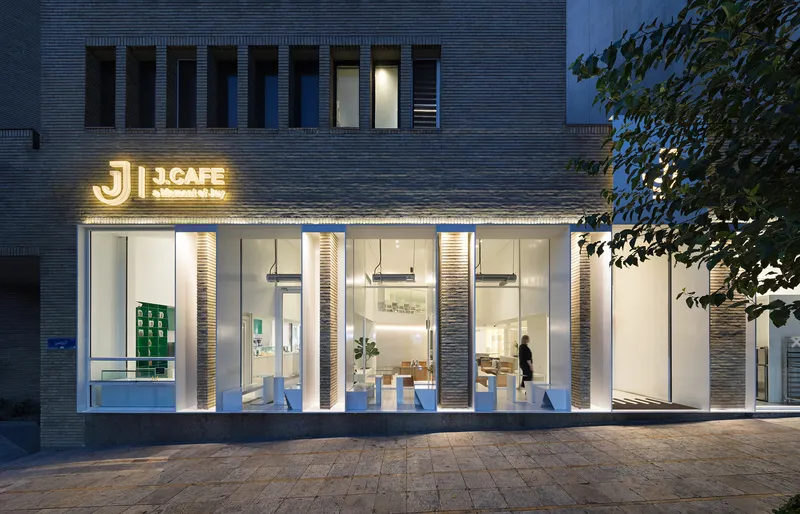 Evening view of the illuminated storefront showing interior shelving and a figure walking through the glass-walled entrance
