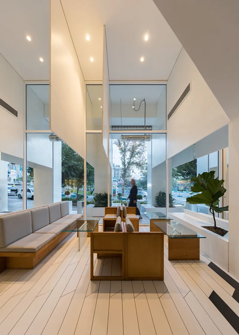 Double-height lobby with timber benches and glass block tables beneath a mezzanine opening with a walking figure silhouetted