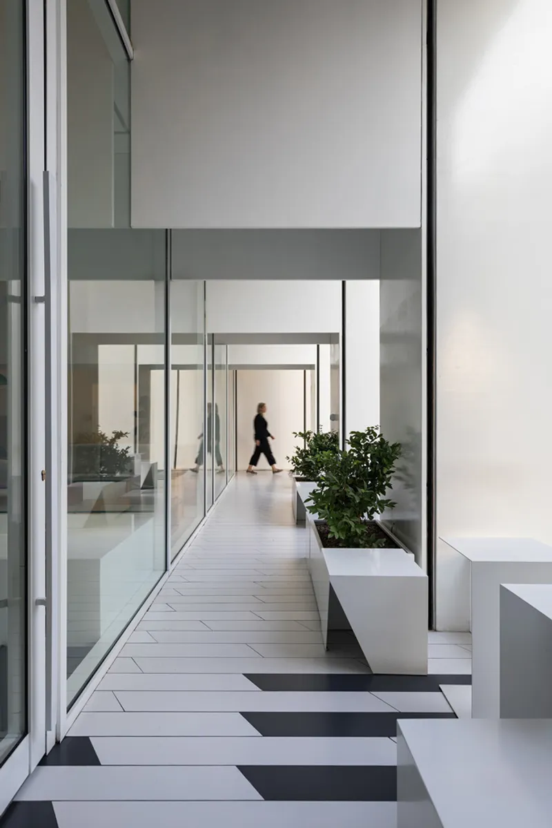 Corridor with floor-to-ceiling glazing and planted white boxes along one edge as a figure walks toward light