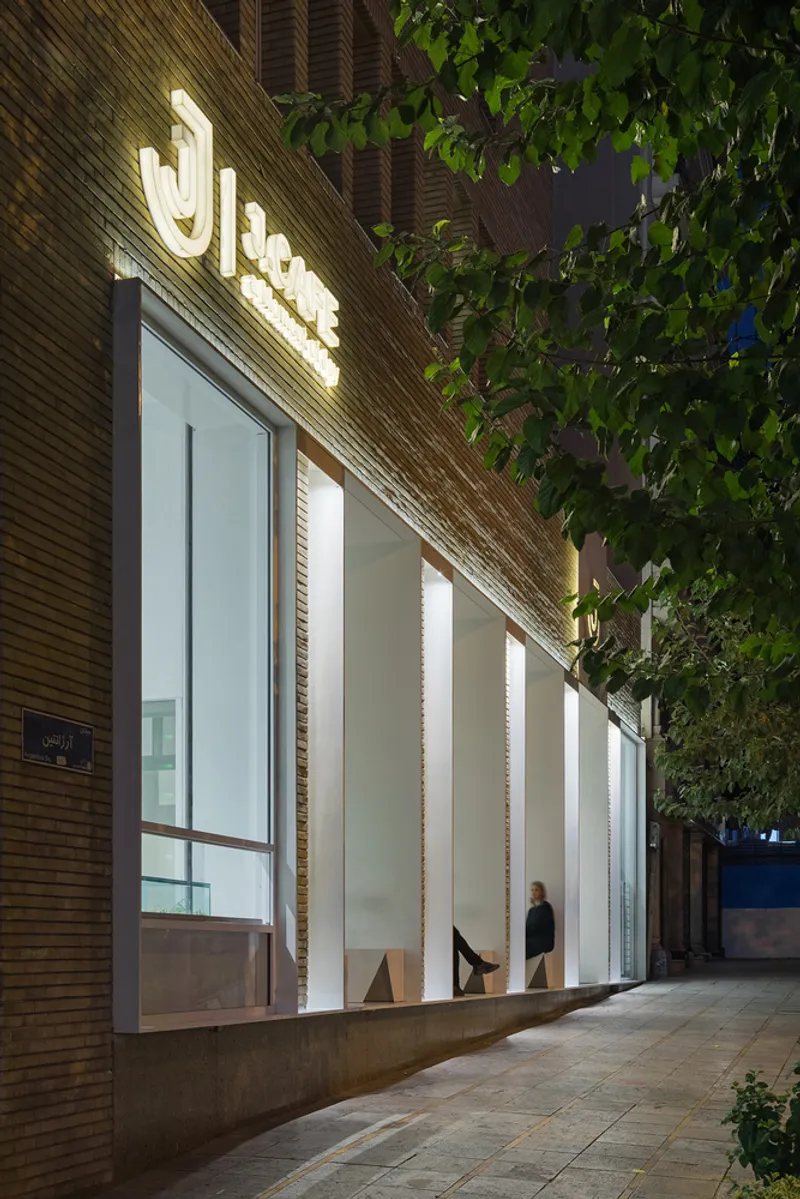 Illuminated storefront facade at dusk with large glass panels and brick cladding beneath tree branches