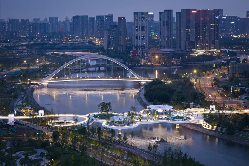 Dusk aerial view of the waterside park with curved white paths and the arched bridge beyond