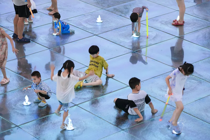 Overhead view of children playing on the shallow water plaza with glass pavers and toy boats