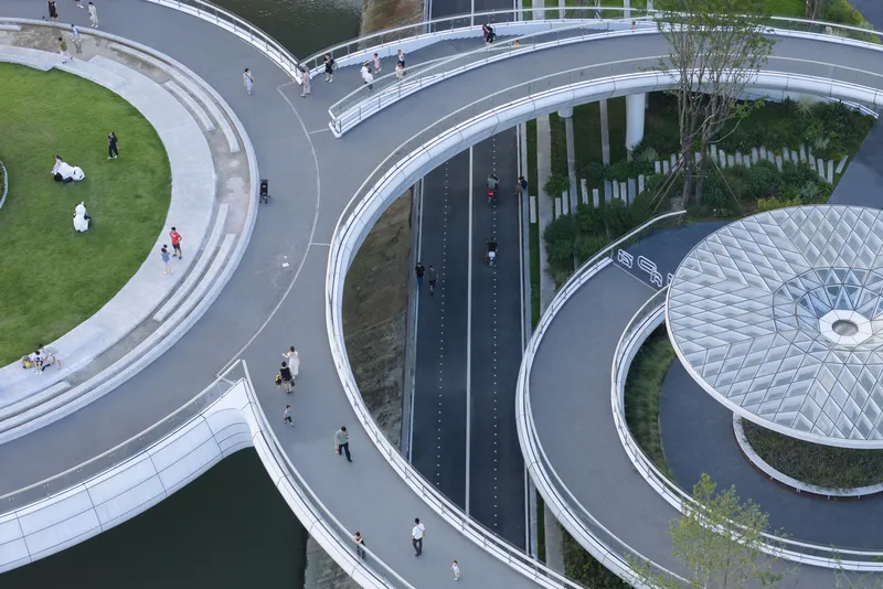 Aerial view of curving pedestrian pathways with circular planted beds and cyclists crossing the bridge deck