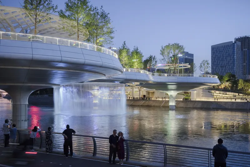 Tiered bridge structure illuminated at dusk with pedestrians gathered along the riverfront promenade watching fountains