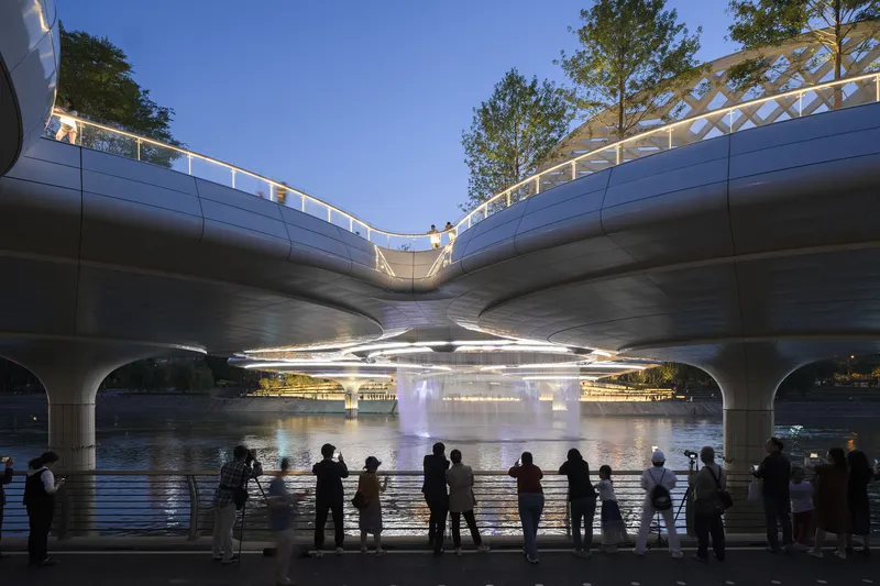 View beneath the curved concrete bridge decks at twilight with crowds gathering along the water's edge