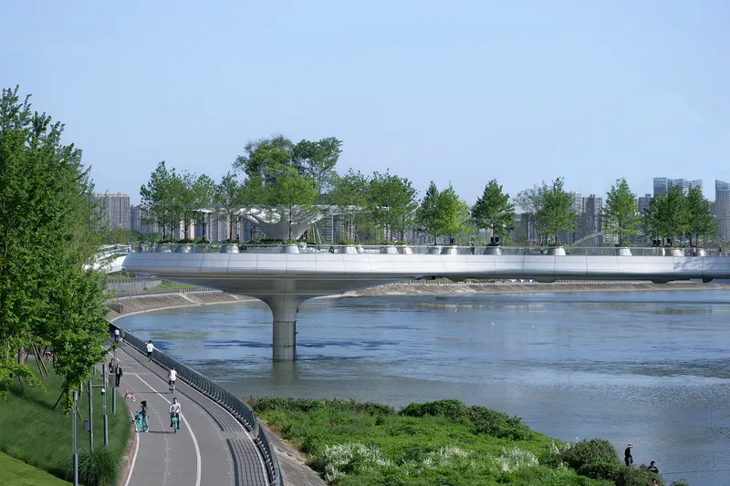 Roadway bridge with planted terrace crossing the river under a cloudy sky with cyclists on the embankment below